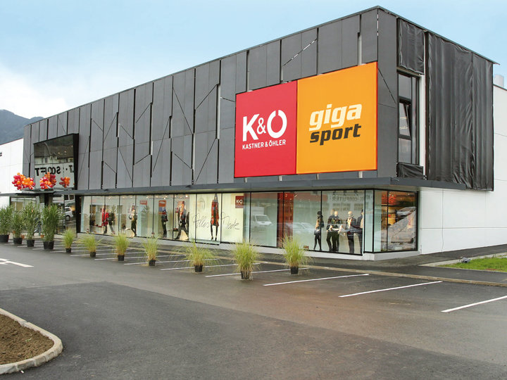 Un immeuble commercial moderne avec un grand panneau rouge-orange indiquant « K&O Kastner & Öhler » et « giga sport ». La façade extérieure est dotée de panneaux sombres et de grandes vitrines où l'on peut voir des mannequins. Au premier plan, on aperçoit un petit parking.