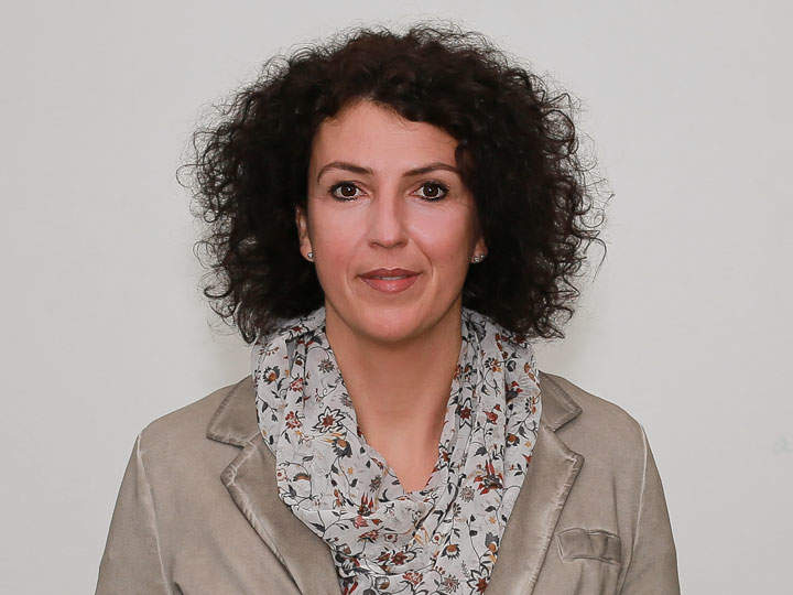 Une personne aux cheveux bouclés et foncés, portant un foulard à fleurs et un blazer beige, regarde directement la caméra. L'arrière-plan est simple et neutre.