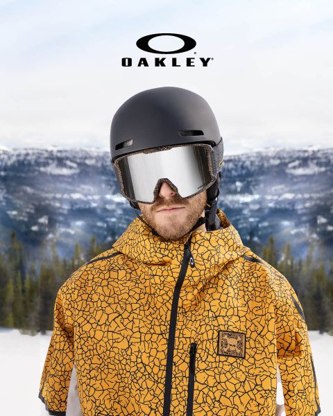 Une personne portant un casque noir, des lunettes de protection réfléchissantes et une veste jaune à motif crépitant se tient devant un paysage montagneux enneigé. Le logo Oakley est visible en haut de l'image.