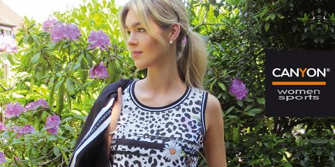 Une femme portant un haut sans manches à motifs noirs et blancs se tient à l'extérieur, à côté de fleurs violettes épanouies et de plantes vertes. Le logo "CANYON women sports" est visible sur le côté droit. Elle tient une veste noire sur son épaule.