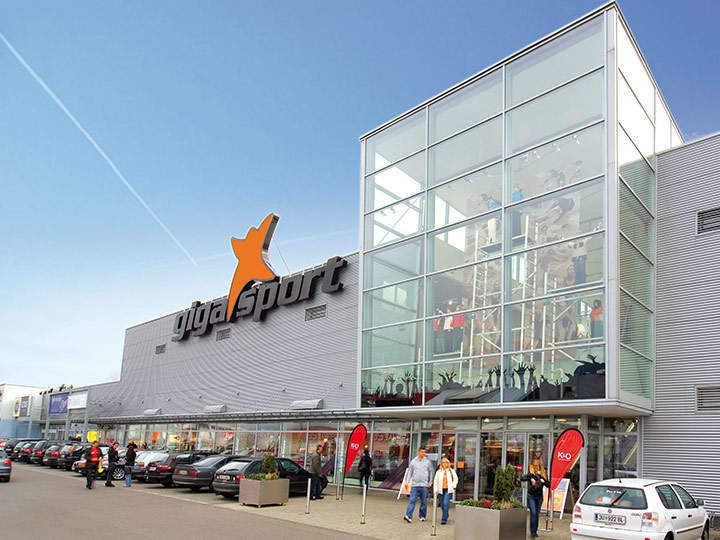 Vue extérieure d'un grand magasin d'articles de sport avec une façade en verre et un logo orange. Plusieurs voitures sont garées sur le parking et quelques personnes se dirigent vers l'entrée. Le bâtiment a un design moderne sous un ciel bleu clair.