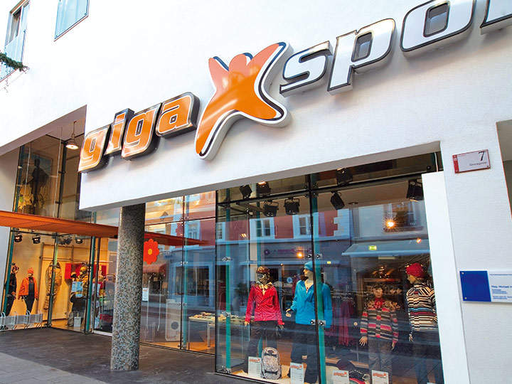 Vue de rue d'un magasin Gigasport avec une façade moderne. L'entrée est flanquée de grandes vitrines où l'on peut voir des mannequins portant des vêtements de sport colorés. Un panneau "Gigasport" bien visible est placé au-dessus de la porte.