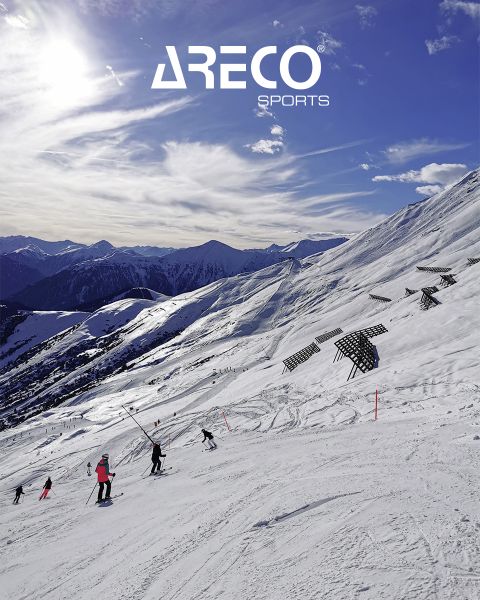 Des skieurs sur une pente de montagne enneigée sous un ciel bleu avec quelques nuages. Le soleil brille intensément et projette des ombres sur la neige. En haut, on peut lire le texte « ARECO SPORTS ». À l'arrière-plan, on aperçoit des sommets enneigés.
