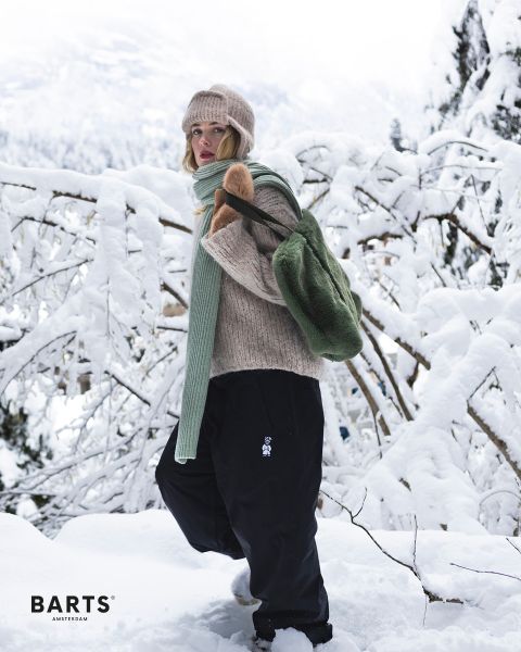 Une personne portant un bonnet en tricot beige, un pull, une écharpe verte et un pantalon noir se tient dans un paysage enneigé et porte un sac à dos vert. Des branches et des arbres enneigés caractérisent l'arrière-plan. Le texte sur l'image est « BARTS AMSTERDAM ».