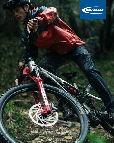 Un cycliste en veste rouge et casque roule sur un sentier accidenté avec son VTT. Le vélo a une fourche rouge et des freins à disque. L'arrière-plan est flou avec du vert. Le logo Schwalbe est visible dans le coin supérieur droit.