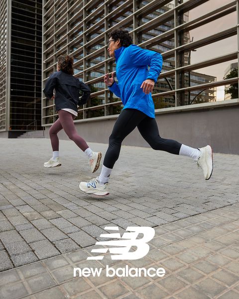 Deux personnes font du jogging à l'extérieur, l'une portant une veste bleue et des leggings noirs, l'autre une veste noire et des leggings marron. Les deux portent des baskets blanches. Elles courent à côté d'un bâtiment moderne avec des panneaux de verre. Le logo New Balance est visible en bas.