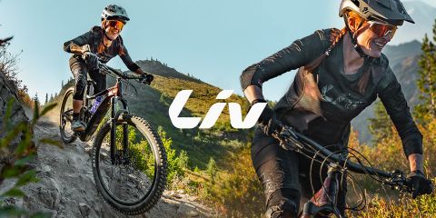 Deux femmes font du VTT sur un sentier accidenté avec des arbres et des montagnes en arrière-plan. Elles portent toutes les deux des casques et des lunettes de soleil. Le mot « Liv » est clairement visible au centre.