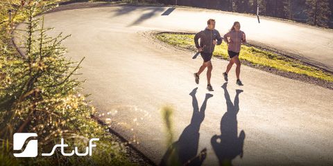 Deux personnes font du jogging sur une route sinueuse entourée d'une forêt. Le soleil projette de longues ombres sur le trottoir. Les deux portent des vêtements de sport. Dans le coin inférieur gauche se trouve un logo avec l'inscription "Stuf".