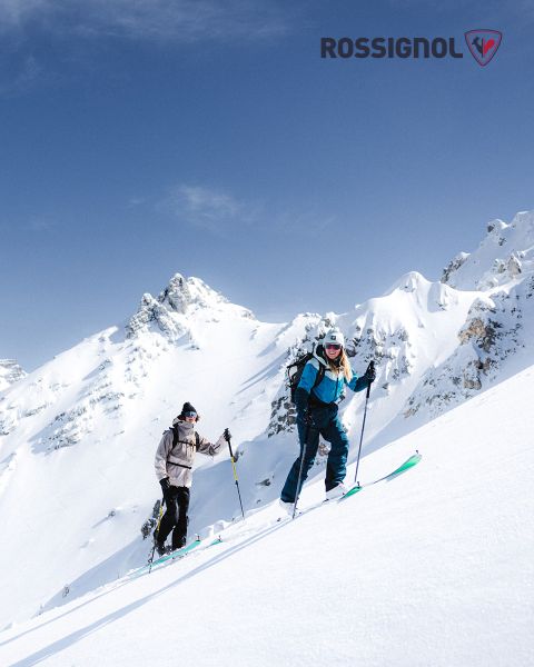 Deux skieurs en tenue d'hiver montent une pente de montagne enneigée par une journée claire. En arrière-plan, on aperçoit des sommets escarpés et enneigés sur un ciel bleu. Le logo Rossignol est visible dans le coin supérieur droit.
