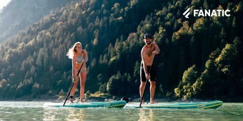 Une femme en bikini et un homme en short et chapeau pagayent sur un lac paisible entouré de montagnes boisées. Ils ont l'air heureux et détendus. Le logo "FANATIC" est visible dans le coin supérieur droit.