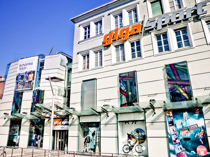 Vue de rue du magasin Gigasport avec de grandes vitrines exposant des articles de sport. Un panneau d'affichage montre des coureurs. Le bâtiment a une façade moderne avec "Gigasport" en lettres orange et grises audacieuses.