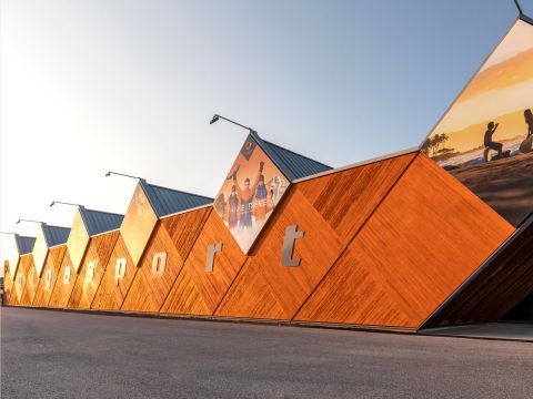 Bâtiment moderne avec une façade hexagonale en bois et l'inscription « ggysport ». Dans la partie supérieure, on peut voir de grandes affiches, dont des images de personnes pratiquant des activités de plein air. Un ciel bleu éclatant est visible en arrière-plan.