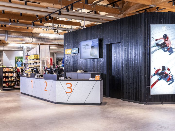 Un intérieur moderne d'un magasin de sport avec un comptoir de service à la clientèle élégant portant les inscriptions 1, 2, 3. Des images sportives sont affichées sur le mur derrière et il y a des étagères bien rangées avec divers articles de sport. Le plafond présente des poutres et des tuyaux apparents.
