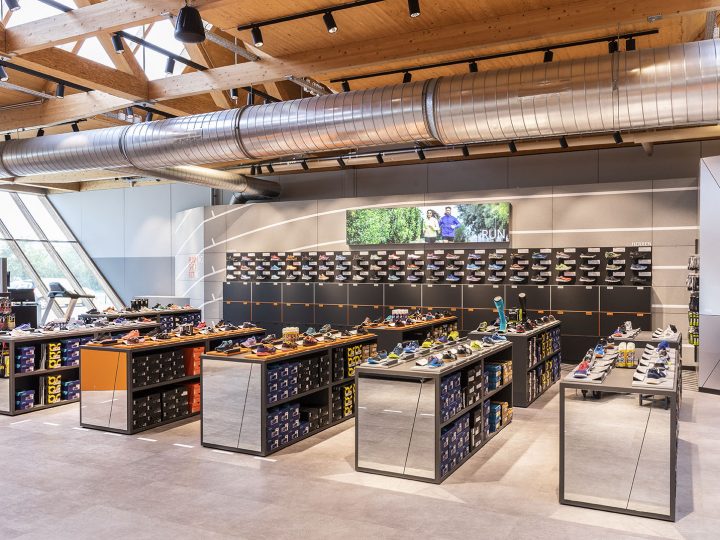 Un magasin de chaussures moderne avec des plafonds en bois, des conduits apparents et de grandes fenêtres. Des présentoirs de chaussures présentant différents styles de chaussures sont répartis dans tout l'espace. Un écran numérique sur le mur du fond affiche une image de personnes à l'extérieur.