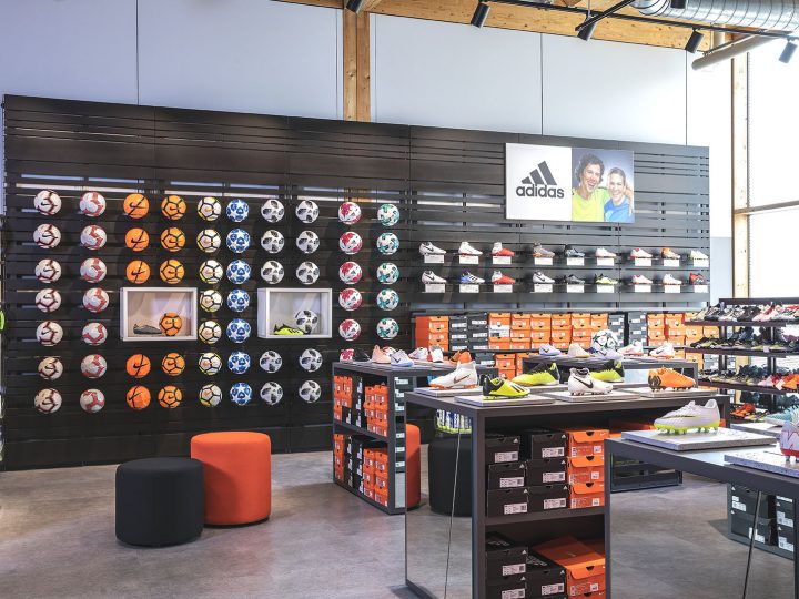 Aménagement intérieur d'un magasin de sport avec une présentation murale de différents ballons de football et des étagères présentant des chaussures Adidas. En dessous, des boîtes à chaussures orange vif sont empilées de manière ordonnée. Le magasin a un design moderne avec des accents de bois et un éclairage industriel.