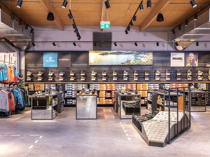 Un magasin de chaussures moderne avec une sélection de chaussures d'extérieur sur des étagères. Les marques sont visibles sur un présentoir mural. Le magasin dispose d'un éclairage élégant, d'accessoires en miroir et d'un espace pour essayer les chaussures sur une rampe texturée. Des vêtements sont situés sur le côté gauche.