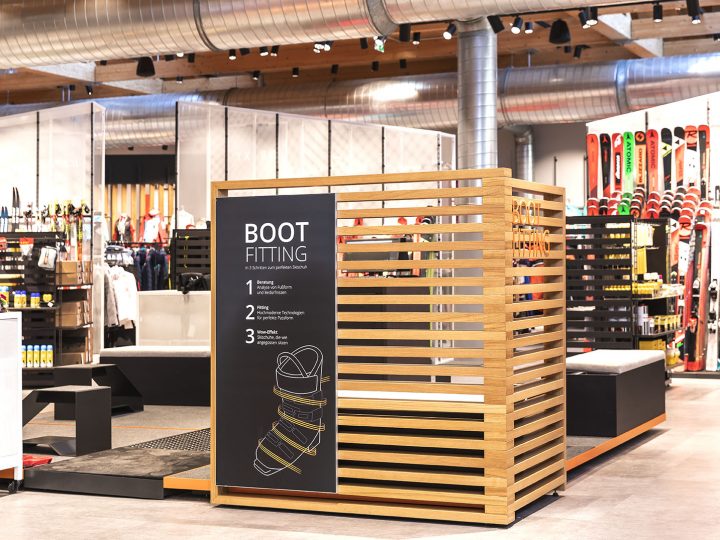 Un espace d'essayage de chaussures dans un magasin de ski, avec un élégant boîtier en bois et un panneau d'indication noir. En arrière-plan, divers équipements de ski, notamment des skis et des bâtons, sont soigneusement disposés sur des étagères.