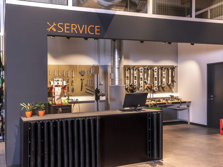 Magasin de service de ski avec un comptoir à l'avant, des outils montés au mur et un poste de travail à l'arrière. À gauche, l'équipement de ski est suspendu à des supports. Le mot « SERVICE » est affiché au-dessus du comptoir.