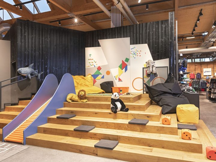 Aire de jeux pour enfants avec des marches en bois, un toboggan bleu et des motifs muraux géométriques colorés. Des animaux en peluche, dont un lion et un panda, ainsi que des poufs sont placés sur les marches. L'espace se trouve dans un environnement industriel moderne avec des tuyaux apparents et des touches de bois.