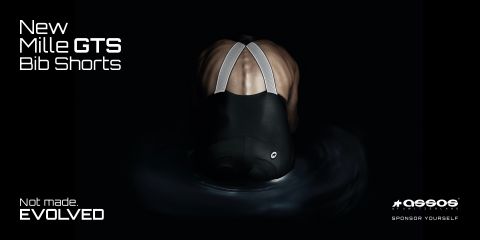 Une personne est vue de dos portant un cuissard de cycliste, avec le texte « New Mille GTS Bib Shorts » dans le coin supérieur gauche. L'image a un fond sombre qui met en valeur les bretelles réfléchissantes et le matériau du cuissard.