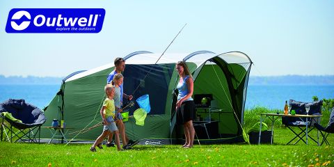 Une famille de quatre personnes profite d'une journée ensoleillée en camping au bord de l'eau. Deux enfants tiennent des épuisettes, tandis que deux adultes se tiennent à côté. Une grande tente verte est installée, entourée de chaises de camping et de matériel. Le logo Outwell est visible dans le coin supérieur gauche.