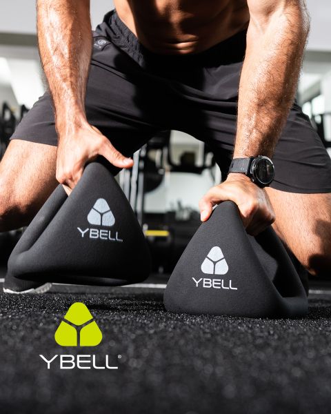 Une personne en tenue de sport tient deux poids YBELL noirs sur le sol de la salle de sport. Elle porte une montre et un short. Le logo YBELL est visible sur les poids et dans le coin inférieur gauche.