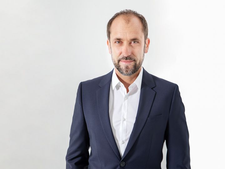 Un homme aux cheveux courts et à la barbe, vêtu d'un costume bleu marine et d'une chemise blanche, se tient devant un fond blanc uni et regarde la caméra avec un léger sourire.