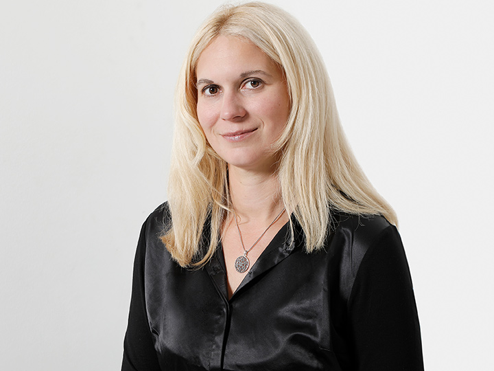 Une personne aux longs cheveux blonds, portant un chemisier noir et un collier avec un pendentif rond, se tient devant un fond blanc uni et sourit légèrement.