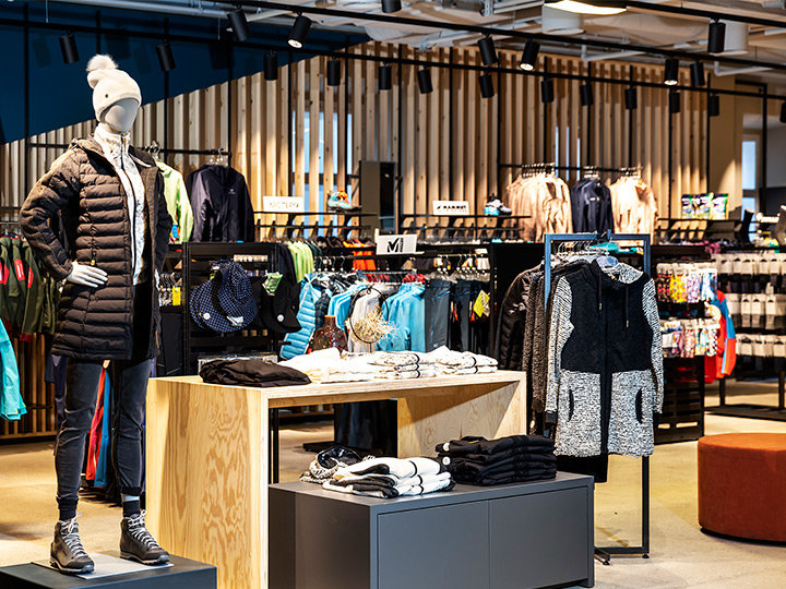 Vue intérieure d'un magasin de vêtements avec un mannequin portant une veste noire, un chapeau blanc et des bottes. Diverses vestes colorées et vêtements de sport sont exposés sur des présentoirs et des étagères, avec des accents de bois et un éclairage moderne en arrière-plan.
