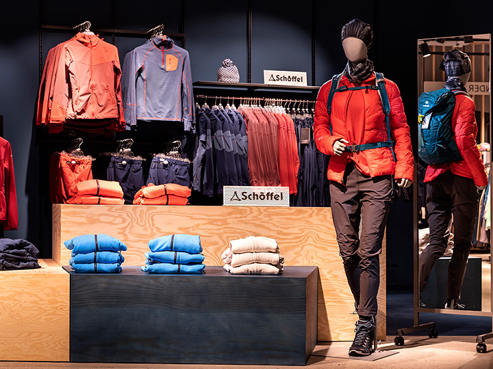 Mannequin en tenue d'extérieur avec une veste orange, un pantalon noir, un sac à dos bleu et un bonnet tricoté. Des vêtements bleus et beiges pliés et des cintres avec des vestes sont disposés sur les étagères d'exposition. Le logo Schöffel est visible en arrière-plan.