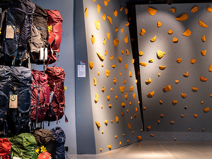 Mur d'escalade intérieur avec différentes prises orange sur un fond gris. À gauche, plusieurs sacs à dos de différentes couleurs sont exposés sur des étagères. L'éclairage est vif et le sol est poli.