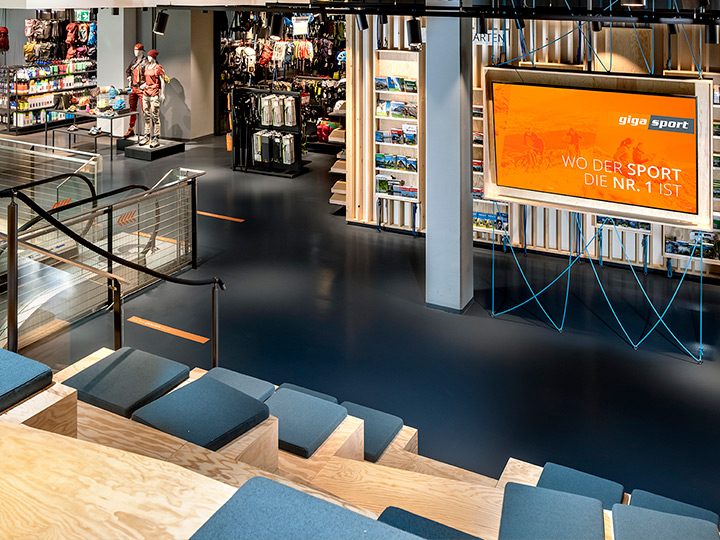 Vue intérieure d'un magasin de sport avec des sièges en bois avec des coussins, un grand écran affichant les publicités "Gigasport" et "Là où le sport est n°1", des escaliers mécaniques et des articles de sport exposés. L'espace est moderne et bien éclairé.