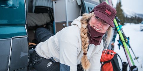 Une personne avec une longue tresse et un bonnet marron souriant en descendant d'une camionnette. Elle est chaudement vêtue, entourée de neige, et on aperçoit des skis et du matériel de ski à l'extérieur.