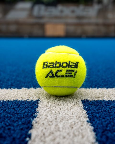 Une balle de tennis Babolat jaune repose sur un court de tennis bleu, centrée sur une ligne blanche. L'arrière-plan est flou, mettant en valeur le logo de la marque et la texture de la balle.