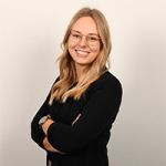 Une personne aux longs cheveux blonds et portant des lunettes sourit, vêtue d'une tenue noire et croisant les bras. L'arrière-plan est simple et clair.
