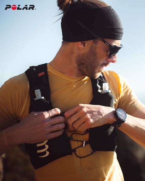 Une personne portant un bandeau noir et des lunettes de soleil ajuste son gilet d'hydratation noir. Elle porte une chemise jaune et une montre intelligente au poignet. Le logo Polar est visible en haut à gauche. La lumière du soleil illumine la scène.