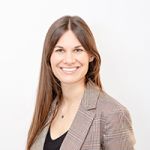 Femme souriante aux longs cheveux bruns, portant un blazer à carreaux et un haut noir, se tenant devant un fond blanc uni.