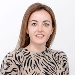Une femme aux cheveux bruns sourit à la caméra. Elle porte un pull beige avec un motif zébré noir. Le fond est uni et blanc.