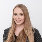 Une personne aux longs cheveux blonds lisses sourit à la caméra. Elle porte une veste en cuir noire et un haut noir. Le fond est simplement blanc.