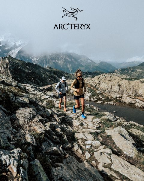 Deux personnes randonnent sur un sentier de montagne rocailleux. Le ciel est couvert, une chaîne de montagnes brumeuse est visible en arrière-plan. Les deux randonneurs portent des équipements de plein air. Un petit étang se trouve à droite. Le logo Arc'teryx est visible en haut.