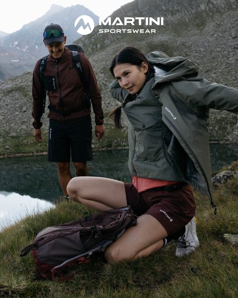 Deux personnes en équipement de plein air avec des sacs à dos se tiennent au bord d'un lac de montagne. L'une est agenouillée et ajuste sa veste, l'autre se tient là en souriant. En arrière-plan, un terrain rocheux et un ciel nuageux. En haut de l'image, on peut lire « MARTINI SPORTSWEAR ».