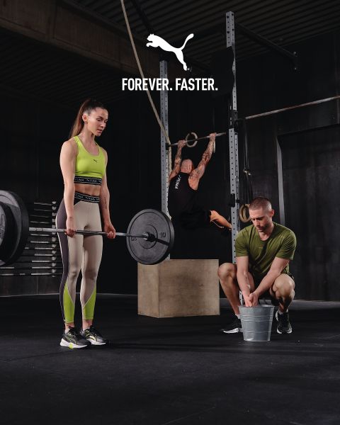 Une femme en tenue de sport soulève un haltère, un homme est accroupi avec un seau à côté d'elle, une autre personne est suspendue à des anneaux dans une salle de sport. En haut, on peut lire le texte « FOREVER. FASTER. » avec un logo Puma.