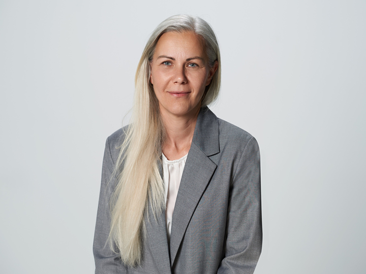 Une personne aux longs cheveux lisses et clairs porte un blazer gris et un chemisier blanc. Elle est assise et regarde la caméra avec une expression neutre devant un fond clair et uni.