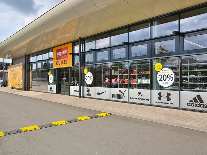 Vue extérieure d'un magasin d'usine d'articles de sport avec de grandes fenêtres affichant divers logos de marques telles que Puma et Adidas. Des panneaux sur les fenêtres annoncent une réduction de 20 % sur les meubles, les rideaux et le linge de maison. Le bâtiment se distingue par son design moderne.