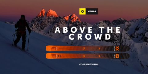 Un skieur se tient au crépuscule sur une montagne enneigée, se détachant comme une silhouette sur le ciel. À l'arrière-plan, on aperçoit les sommets illuminés. Au premier plan, on voit des skis Völkl orange. Le texte est « Above the Crowd » avec les hashtags #THISISSKTOURING.