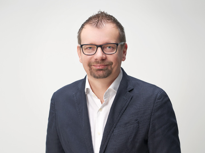 Un homme aux cheveux courts et portant des lunettes arbore un blazer bleu marine sur une chemise blanche. Il sourit et se tient devant un fond clair et uni.