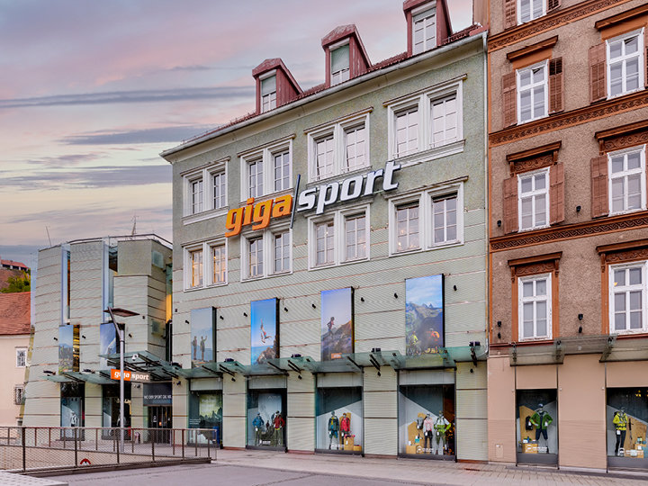 Vue de rue d'un magasin Gigasport avec une façade vert clair et de grandes fenêtres présentant des articles de sport de plein air. L'entrée est dotée d'un toit en verre moderne et le bâtiment est adjacent à un bâtiment en briques brunes. Le ciel est nuageux et laisse entrevoir un coucher de soleil.