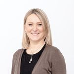 Une femme aux cheveux blonds mi-longs et souriante porte un haut noir et un cardigan marron. Elle porte un collier de perles et se tient devant un fond blanc uni.
