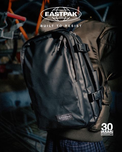 Une personne portant une veste marron porte un sac à dos Eastpak noir avec le slogan "Built to Resist". L'image montre un logo et la mention d'une "garantie de 30 ans". L'arrière-plan est flou.