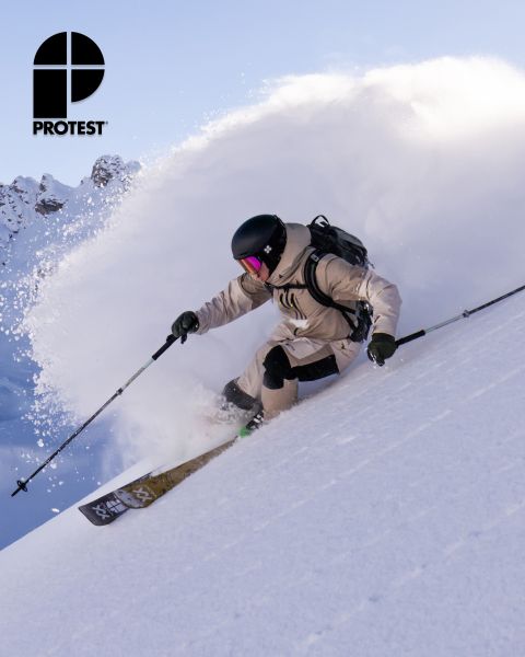 Un skieur en équipement beige et casque dévale une pente raide dans la neige fraîche, laissant derrière lui un nuage de neige. Le logo « PROTEST » est visible dans le coin supérieur gauche. En arrière-plan, on aperçoit un ciel clair et des montagnes enneigées.